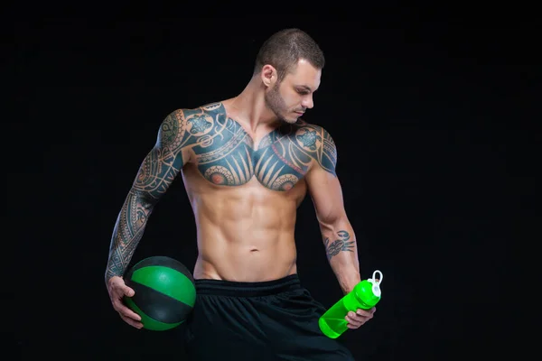 Gespierde man bodybuilder met tatoeages. Man poseren op een zwarte achtergrond met water fles en bal — Stockfoto