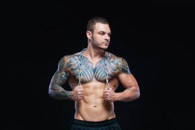 Dövmeli güçlü kaslı atletik adam siyah arka planda boynuna metal zincir takıyor.