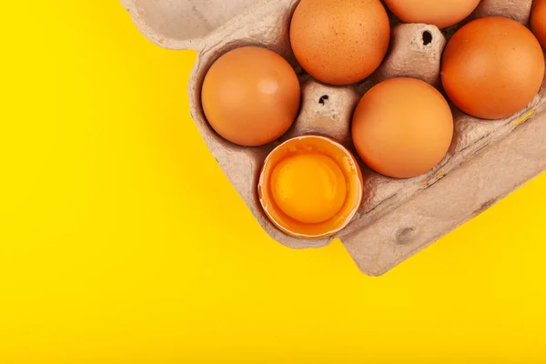 Eieren Kippeneieren. Bovenaanzicht van een open grijze doos met bruine eieren geïsoleerd op een gele achtergrond. Eén ei is half gebroken, gele ronde dooier. Het concept van een gezonde levensstijl, het krijgen van pure eiwitten — Stockfoto