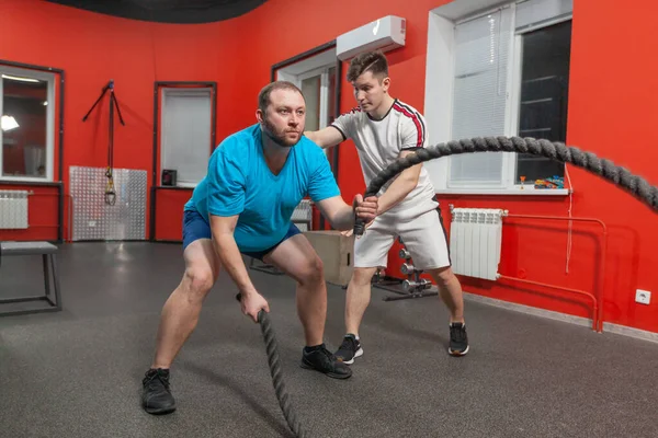 O jovem gordo está muito concentrado realizando exercícios de corda de batalha no ginásio de fitness sob controle de seu treinador pessoal. Peso em excesso — Fotografia de Stock