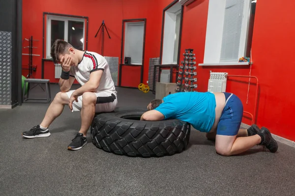 stock image Personal trainer is dissatisfied with his overweight sportsman who is lying exhausted with his head into a tire after hard training in the gym