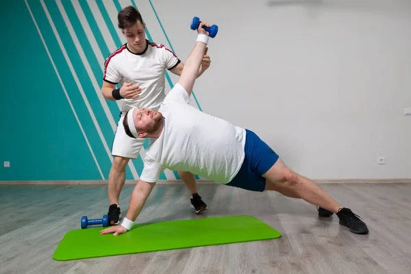Gordo está trabalhando duro no ginásio de fitness com seu personal trainer fazendo exercícios com halteres no limite de possibilidades . — Fotografia de Stock
