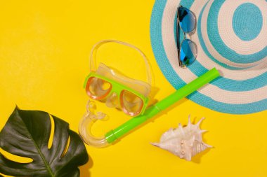 Sarı arkaplanda plaj aksesuarları - güneş gözlükleri, çizgili mavi şapka, dalış maskesi, deniz kabuğu ve canavar yaprağı. Uzun zamandır beklenen deniz ve seyahat tatili kavramı. Reklam alanı