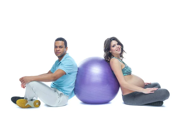 妊娠の最後の数ヶ月の若いカップルとその妻は、ボールの近くのスポーツマットに座っています。黒人と白人の女性一緒に準備する異なるレースの幸せなカップルの概念 — ストック写真