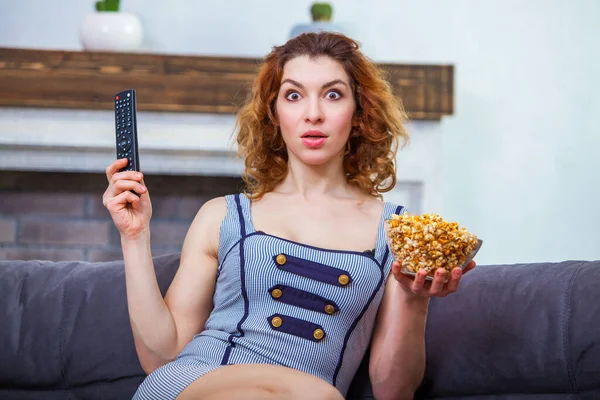 Jeune femme tenant télécommande TV dans une main et un bol de maïs soufflé dans une autre semble très surpris — Photo