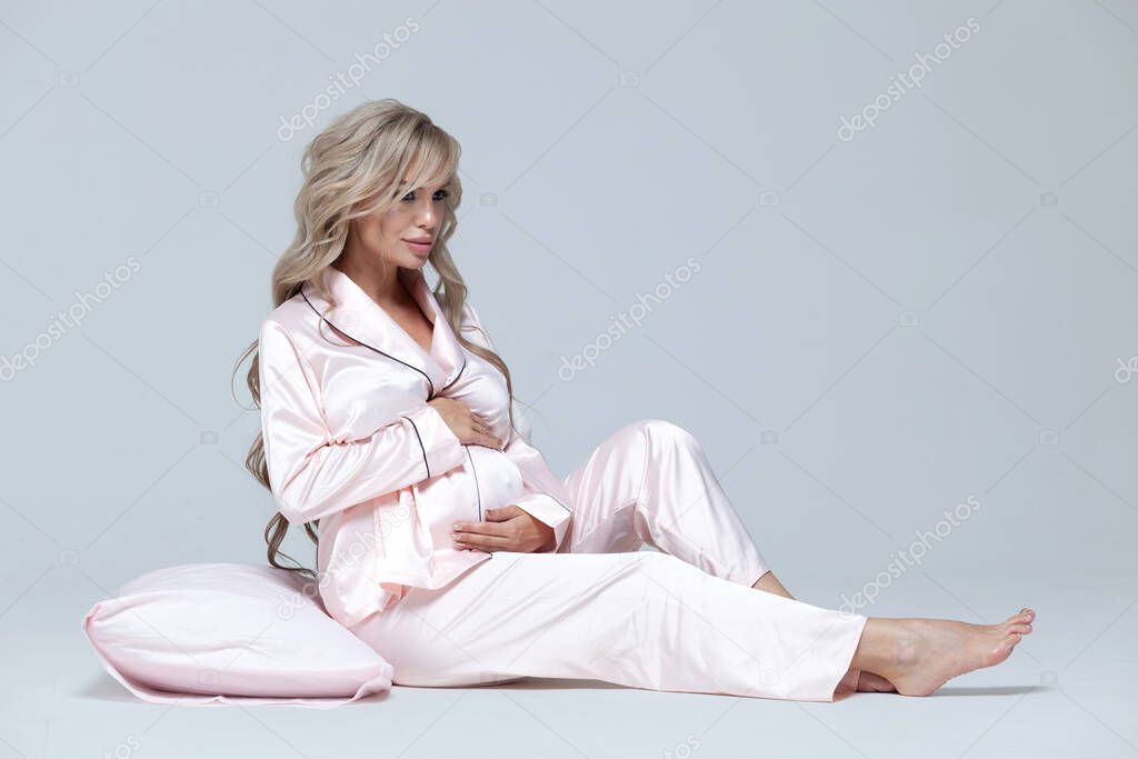 Young beautiful pregnant woman in pink pajamas with pink pillow. Sits and hugs a pillow. The concept of stylish pregnancy and beautiful sleep. On a gray uniform background. Copy paste.