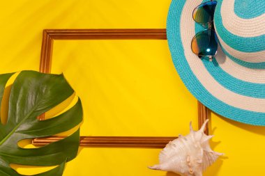 Çizgili mavi şapka, güneş gözlüğü, canavar yaprağı, deniz kabuğu ve sarı arka planda tatil fotoğrafları için çerçeve. Plaj tatili kavramı, deniz turu, sıcak güneşli yaz. Kopyala-yapıştır
