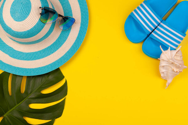 Top view of blue beach accessories on yellow background - sunglasses, flip-flops and striped hat, shell and monstera leaf. Concept of beach holiday, sea tour, warm sunny summer. Advertising space