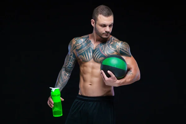 Muskulös man kroppsbyggare med tatueringar. Man poserar på en svart bakgrund med vattenflaska och boll — Stockfoto
