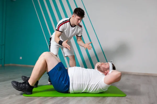 在私人教练的严格控制下，胖男人躺在健身房的垫子上做腹部肌肉运动时，看起来筋疲力尽。体重超标 — 图库照片