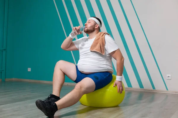 Homem com excesso de peso está sentado em uma bola de fitness exausto após um treinamento duro em aulas de fitness em grupo. Gordo bebe água com uma toalha no ombro. — Fotografia de Stock