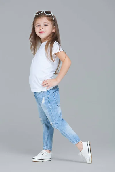Niña preescolar en jeans azules, camiseta blanca y gafas de sol posando aislada sobre fondo gris — Foto de Stock