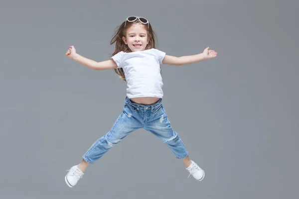 Niña preescolar en vaqueros azules, camiseta blanca y gafas de sol está saltando con las manos levantadas aisladas sobre fondo gris — Foto de Stock