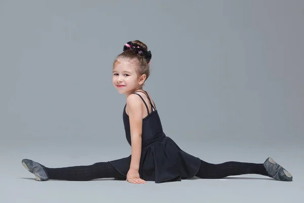 Bela ginasta em sportswear preto mostra exercícios em fundo cinza. Menina flexível fazendo as divisões para se tornar dançarina de balé — Fotografia de Stock