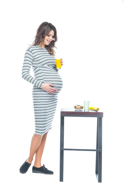 Egy fiatal terhes nő ül egy asztalhoz, amelyen fekszik a banán, van egy tányér gabona, egy pohár kefir és gyümölcslé. A terhesség alatti táplálkozás fogalma. Fehér alapon izolált. — Stock Fotó