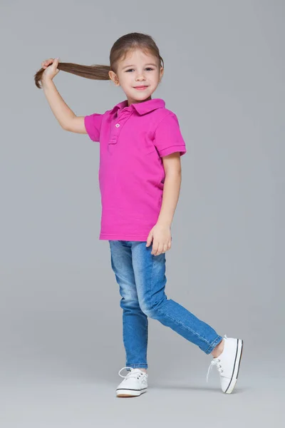 Full length portrait of funny active little girl wearing casual clothes: jeans and pink t-shirt, holding her ponytail in a hand isolated on grey studio background — 스톡 사진