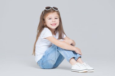 Kid girl preschooler in blue jeans, white t-shirt and sunglasses is posing sitting isolated on grey background