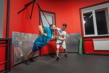 Fat man in a gym is diligently doing pull-ups under the guidance of a personal trainer. Overweight TRX