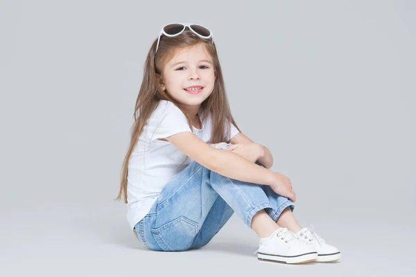 Niña preescolar en jeans azules, camiseta blanca y gafas de sol posando sentada aislada sobre fondo gris — Foto de Stock