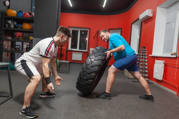 Hombre gordo es muy persistente haciendo ejercicio de volteo de neumáticos en el gimnasio bajo la guía de entrenador personal. Sobrepeso —  Fotos de Stock