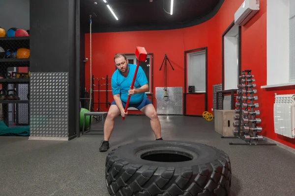 胖男人是一个非常坚持不懈的训练与大锤和轮胎在健身房。体重超标 — 图库照片