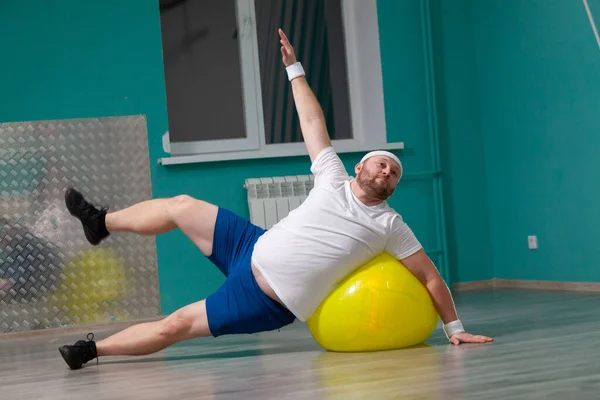 Hombre gordo sonriente está haciendo ejercicios usando la pelota de fitness. Hombre con sobrepeso está contento con el resultado de su entrenamiento en clases de fitness en grupo — Foto de Stock