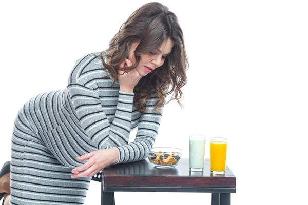 A young pregnant woman sits at a table on which lies a banana, there is a plate with cereal, a glass with kefir and juice. The concept of nutrition during pregnancy. Isolated on a white background. — Stock Photo, Image