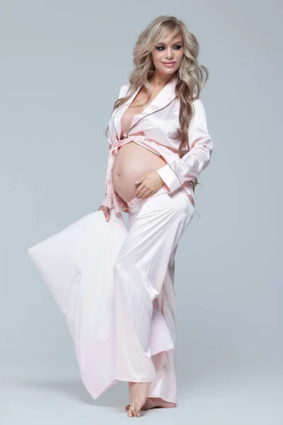 Jeune belle femme enceinte en pyjama rose avec oreiller rose. Le concept de grossesse élégante et de sommeil magnifique. Sur un fond uniforme gris. Copier la pâte. Longueur totale . — Photo