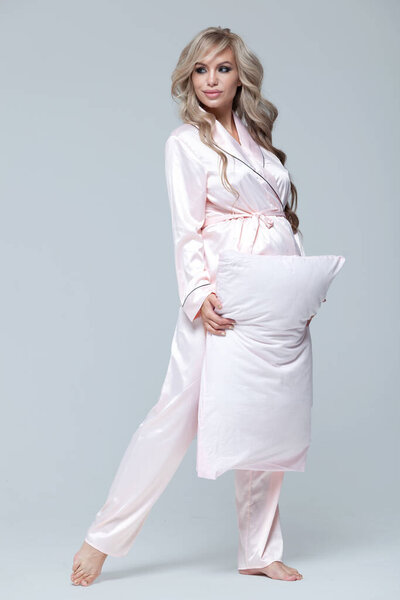 Young beautiful pregnant woman in pink pajamas with pink pillow. The concept of stylish pregnancy and beautiful sleep. On a gray uniform background. Copy paste. Full length.