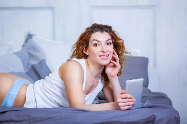 Retrato de uma menina feliz em um top branco deitado na cama com smartphone olhando para a câmera — Fotografia de Stock