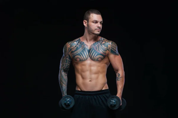 Татуйований сильний м'язовий атлетичний чоловік накачує м'язи гантелями на чорному тлі — стокове фото
