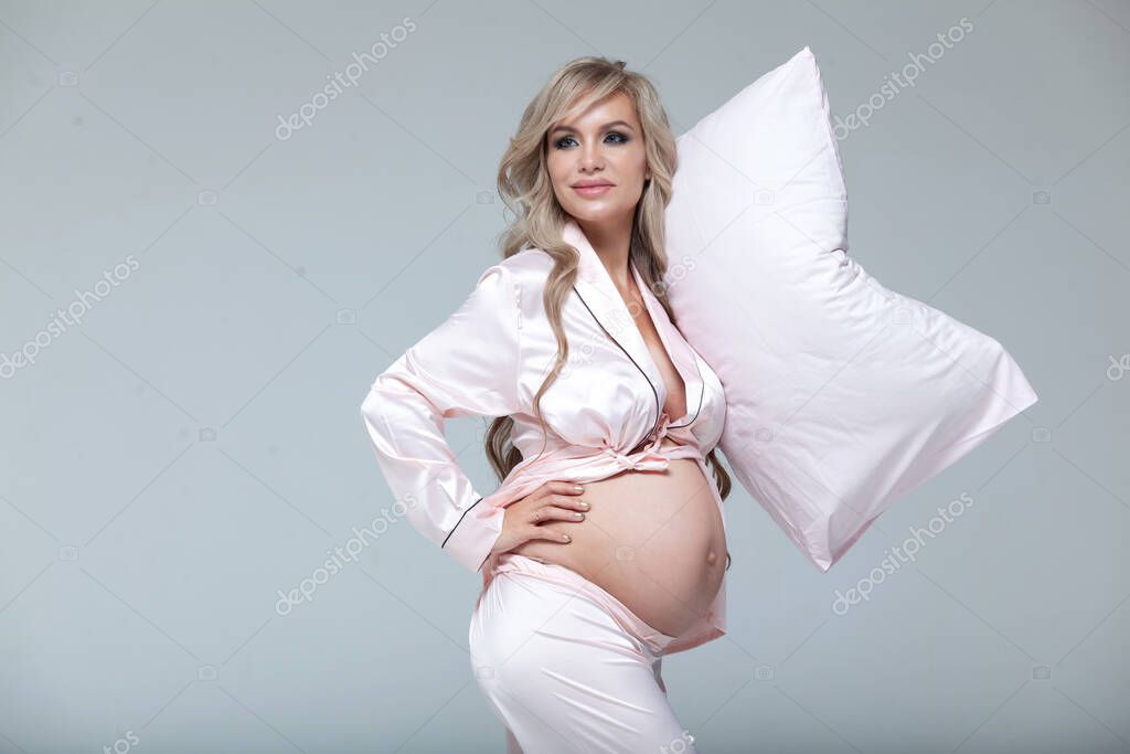 Young beautiful pregnant woman in pink pajamas with pink pillow. The concept of stylish pregnancy and beautiful sleep. On a gray uniform background. Copy paste.