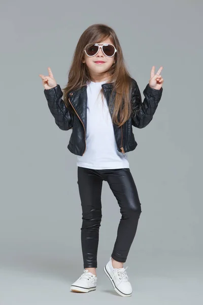 Niña con pelo largo en chaqueta de cuero negro y leggings posa en el estudio con gafas de sol modernas sobre fondo gris — Foto de Stock