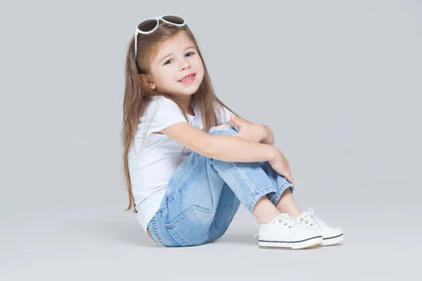 Niña preescolar en jeans azules, camiseta blanca y gafas de sol posando sentada aislada sobre fondo gris — Foto de Stock