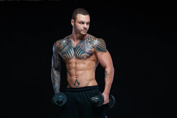 Hombre atlético musculoso fuerte tatuado bombeando músculos con mancuernas sobre fondo negro —  Fotos de Stock