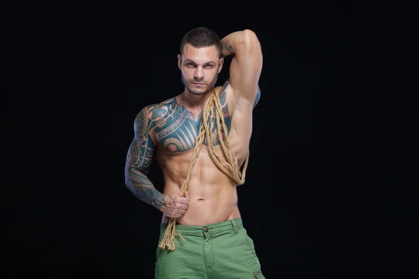 М'язовий чоловік культурист з татуюваннями. Чоловік позує на чорному тлі з мотузкою — стокове фото