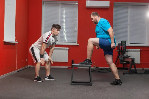 O homem gordo está diligentemente fazendo exercício de intensificação sob o controle de um instrutor pessoal. Sobrepeso — Fotografia de Stock
