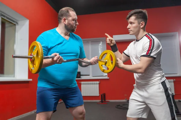健身房里的一个胖子在私人教练的指导下，在他的能力范围内进行杠铃举重练习。体重超标 — 图库照片