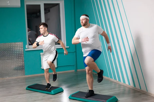Tłuścioch bardzo ciężko pracuje wykonując ćwiczenia step-up na krokach podczas grupowych zajęć fitness. Nadwaga — Zdjęcie stockowe