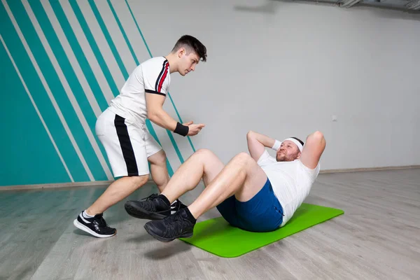 O homem gordo parece exausto ao fazer exercícios nos músculos abdominais deitados no tapete no ginásio de fitness sob controle rigoroso do instrutor pessoal. Sobrepeso — Fotografia de Stock