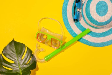 Sarı arkaplanda plaj aksesuarları - güneş gözlükleri, çizgili mavi şapka, dalış maskesi ve canavar yaprağı. Uzun zamandır beklenen deniz ve seyahat tatili kavramı. Reklam alanı