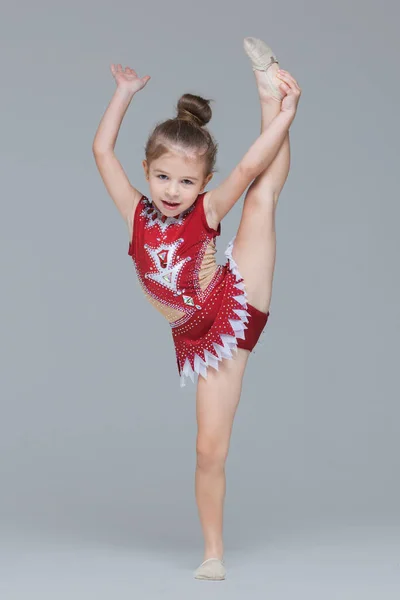 Flexible little girl in beautiful red dress is stretching and doing the splits while training gymnastics on grey background