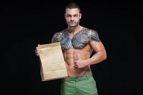 Muscular hombre culturista con tatuajes. Hombre posando sobre un fondo negro con bolsa de papel. El concepto de la publicidad de la comida deportiva adecuada. Copiar logo de pasta . —  Fotos de Stock