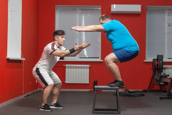 Ansioso homem com excesso de peso está fazendo saltar no ginásio sob a orientação de um personal trainer — Fotografia de Stock