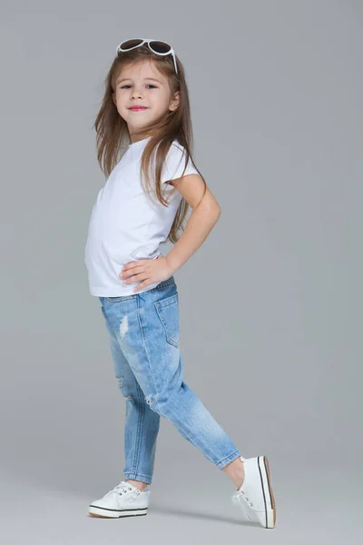 Kid girl preschooler in blue jeans, white t-shirt and sunglasses is posing isolated on grey background — Stock Photo, Image