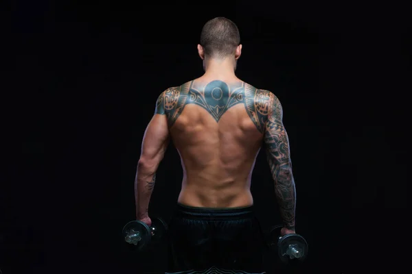 Achteraanzicht van getatoeëerde sterke gespierde atletische man pompen spieren met halters op zwarte achtergrond — Stockfoto