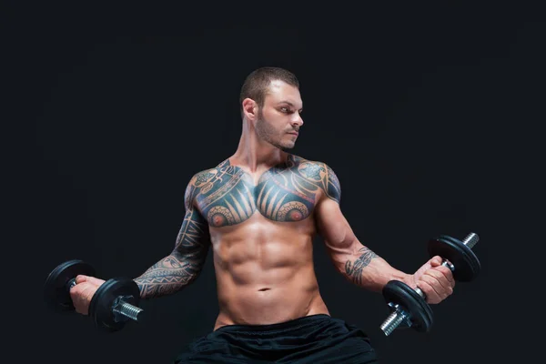 Татуированный сильный мускулистый спортсмен поднимает гантели на черном фоне — стоковое фото