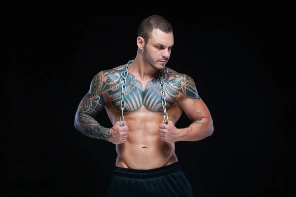 Hombre atlético musculoso fuerte tatuado sosteniendo cadena de metal sobre su cuello sobre fondo negro —  Fotos de Stock