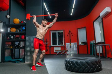 Modern spor salonunda balyozla egzersiz yapan güçlü kaslı bir adamın tam boy portresi.