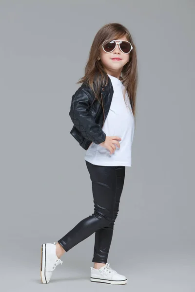 Niña con pelo largo en chaqueta de cuero negro y leggings posa en el estudio con gafas de sol modernas sobre fondo gris — Foto de Stock